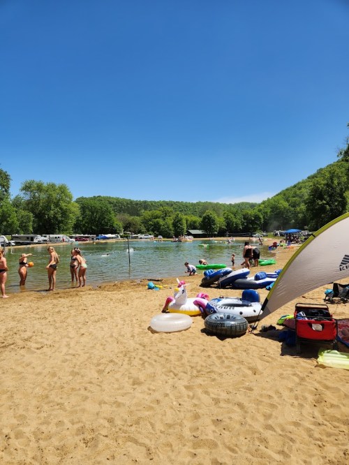 A sunny beach scene with people swimming and relaxing by a lake, surrounded by trees and colorful inflatable floats.