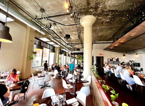 A spacious restaurant interior with diners at tables, exposed beams, and plants hanging from the ceiling.