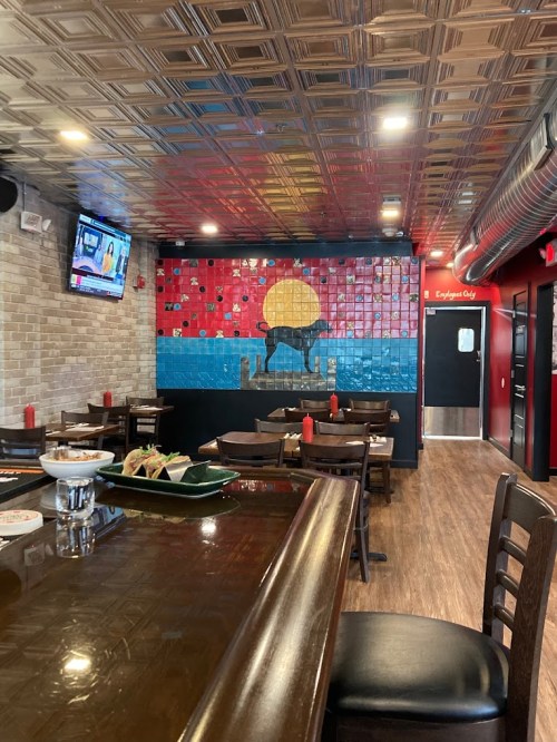 Interior of a restaurant featuring a colorful mural, wooden tables, and a bar area with a TV in the background.