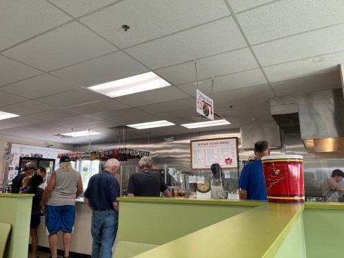 A busy diner with customers in line at the counter, a menu displayed, and staff preparing food in the kitchen.