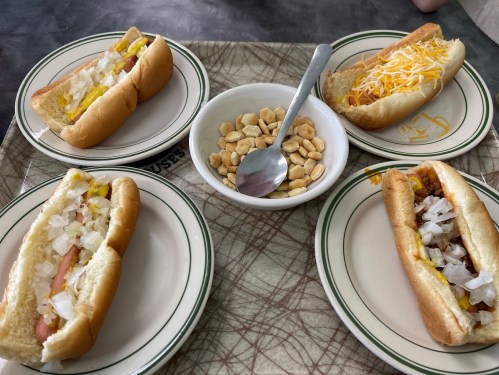 A tray with four hot dogs on plates, topped with onions and mustard, alongside a bowl of peanuts and a spoon.