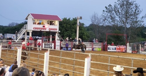 A rodeo scene with cowboys riding horses, spectators watching, and a wooden announcer's booth in the background.