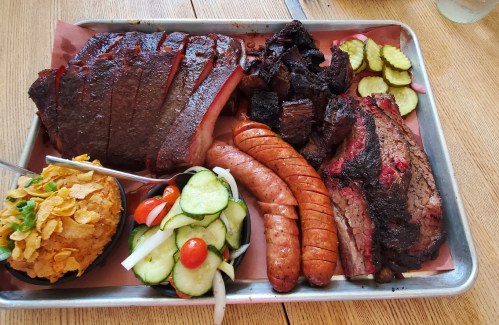 A metal tray filled with various meats, including brisket, sausage, and smoked beef, alongside pickles and a salad.