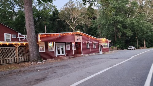 A rustic red building with string lights, surrounded by trees, along a quiet road.