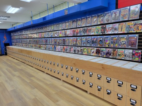 A comic book store interior featuring a wall of comic book displays and wooden storage drawers.
