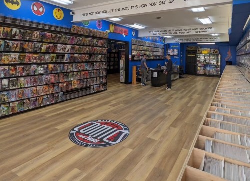 A comic book store interior with shelves of comics, a logo on the floor, and two customers browsing.