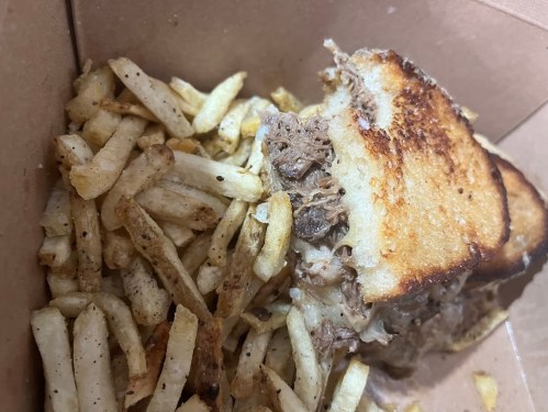 A partially eaten sandwich with pulled meat, served with a side of crispy fries in a takeout box.