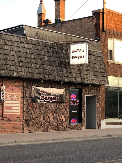 Exterior of Bump's Burgers, featuring a stone facade and a shingled roof, with a sign and neon lights.