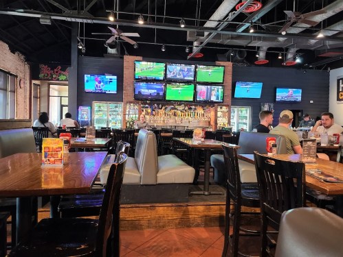 A lively restaurant interior with multiple TVs showing sports, wooden tables, and patrons enjoying their meals.