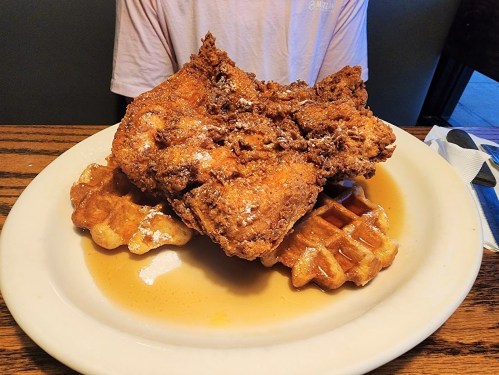 A large piece of fried chicken atop two waffles, drizzled with syrup, served on a white plate.