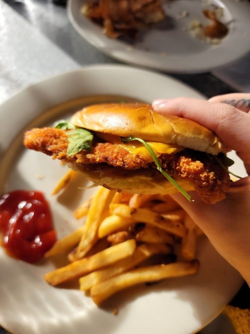 A hand holding a crispy chicken sandwich with lettuce, alongside a plate of fries and ketchup.