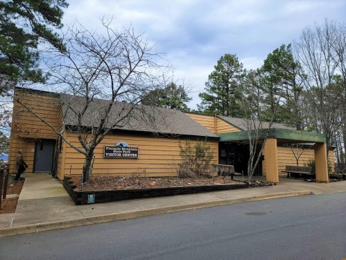A modern visitor center with wooden siding, surrounded by trees and a paved walkway. Cloudy sky in the background.
