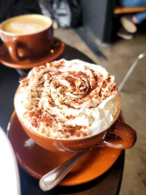 A close-up of a coffee cup topped with whipped cream and cocoa powder, sitting on a dark table.