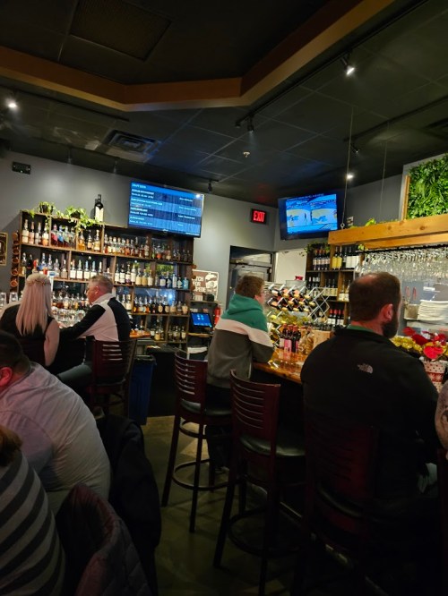 A cozy bar scene with patrons seated at the counter, shelves of liquor, and TVs displaying sports in the background.