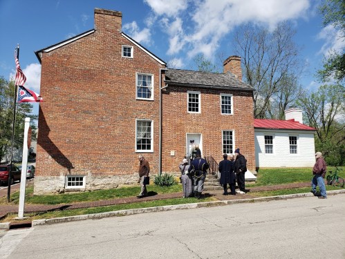 A historic brick building with a red roof, surrounded by people in period clothing, on a sunny day.