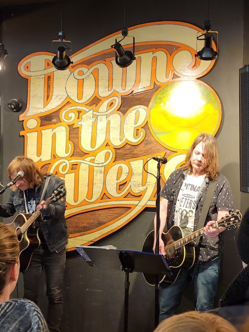 Two musicians perform with acoustic guitars in front of a decorative wall sign that reads "Downs in the Way."
