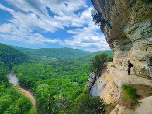 A person stands on a rocky ledge overlooking a lush green valley and river under a partly cloudy sky.