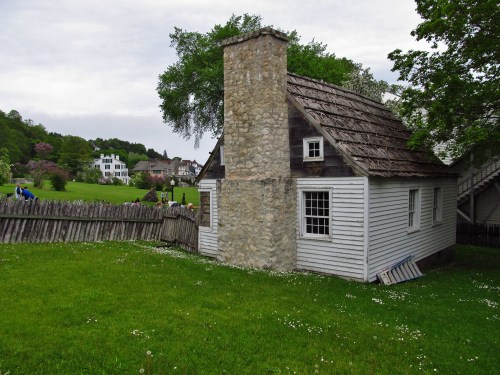 A small, historic house with a stone chimney, surrounded by green grass and trees, in a scenic outdoor setting.