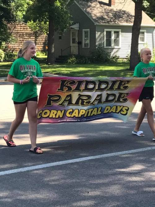 Two girls in green shirts carry a colorful banner that reads "Kiddie Parade Corn Capital Days" during a parade.