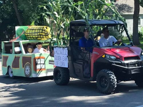 A decorated vehicle with children, resembling a corn-themed float, drives down a street during a parade.