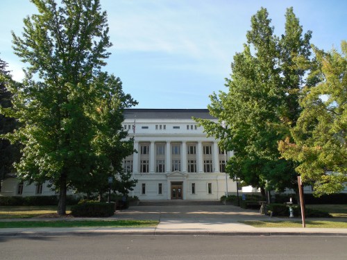 A large, classical-style building with columns, framed by green trees and a clear blue sky.