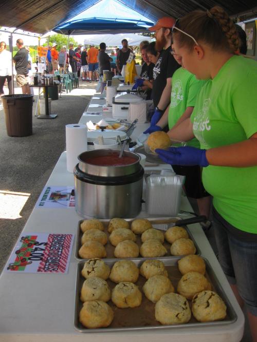 A food booth with volunteers serving dishes, featuring trays of baked goods and a pot of sauce under a blue canopy.
