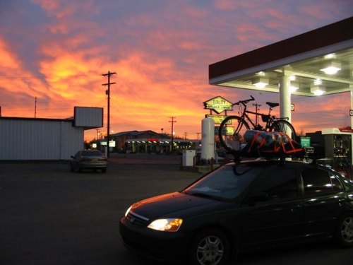 A car with bikes on the roof rack parked at a gas station, with a vibrant sunset in the background.