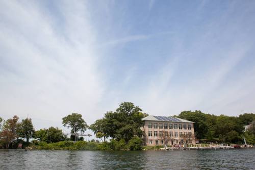 A waterfront view of a modern building surrounded by trees under a blue sky with wispy clouds.