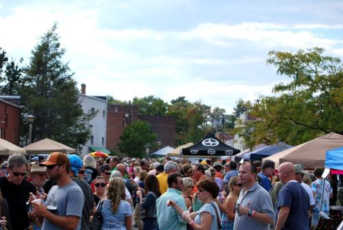A bustling outdoor market scene with a large crowd, tents, and vendors under a partly cloudy sky.
