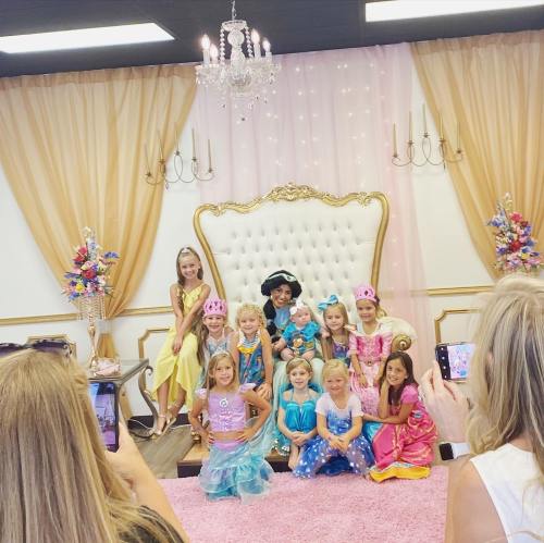 A group of young girls in colorful princess costumes pose together in a decorated room with a large throne.