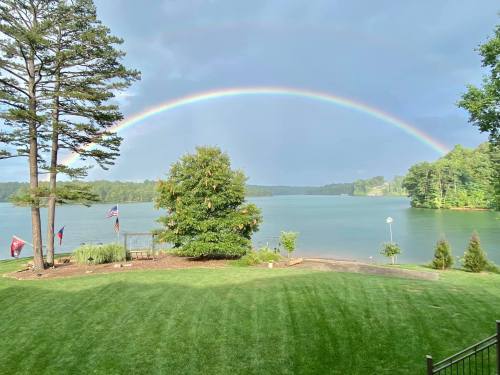 A vibrant rainbow arcs over a serene lake, framed by trees and flags, with a lush green lawn in the foreground.