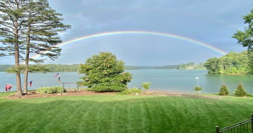 A serene lake view with a vibrant rainbow arching over the water, surrounded by trees and a well-kept lawn.