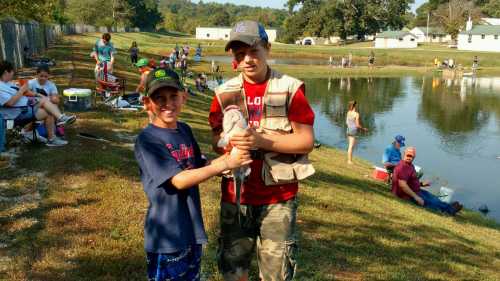 Two boys proudly hold a fish by a pond, surrounded by people fishing and enjoying a sunny day outdoors.