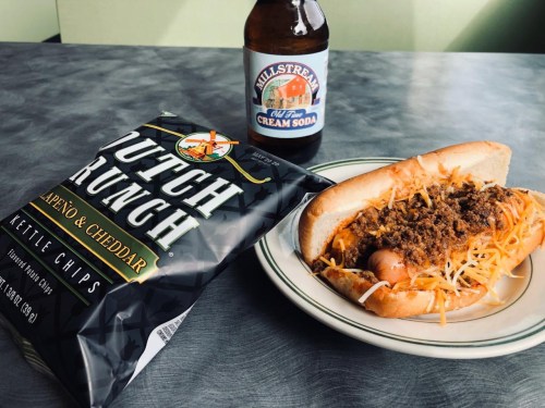 A plate with a chili dog topped with cheese, a bag of kettle chips, and a bottle of cream soda on a table.
