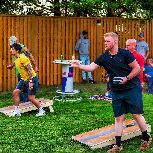 A group of people playing a lawn game, with two players throwing bean bags and others watching in a grassy area.