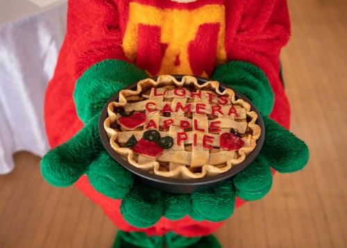 A person in a red and green costume holds a pie decorated with the words "Lights, Camera, Apple Pie."