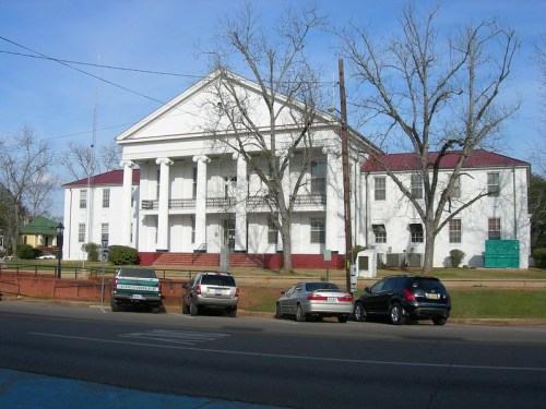 A white building with columns and a red roof, surrounded by trees and parked cars on a sunny day.