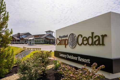 Sign for Camp Cedar, a luxury outdoor resort, with buildings and landscaping in the background.