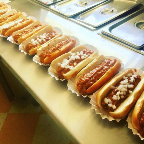 A row of hot dogs topped with chili and diced onions, arranged neatly on a countertop.
