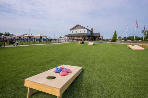 A grassy area with cornhole boards and colorful bags, featuring a building in the background and flags flying.