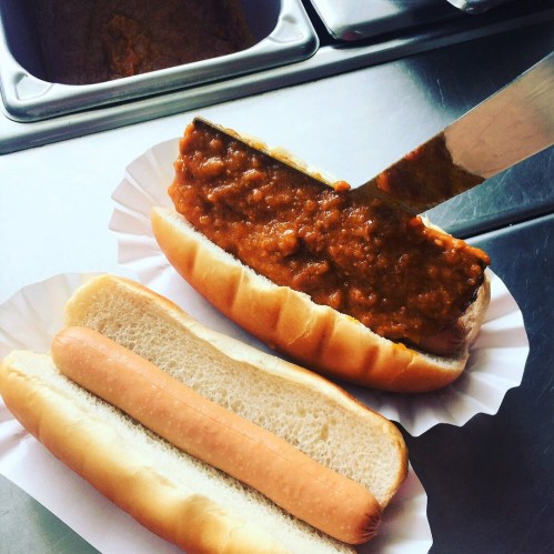 A hot dog bun filled with chili, next to an empty bun and a hot dog on a stainless steel surface.