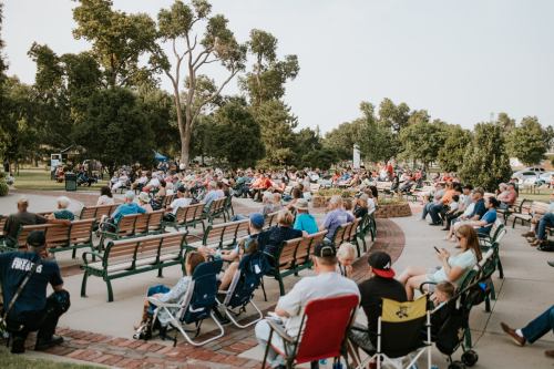 A large crowd of people seated on benches and chairs in a park, enjoying an outdoor event on a sunny day.