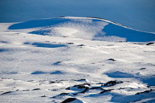 A snowy landscape with gentle slopes and rocky patches under a clear blue sky.