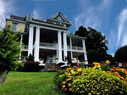 A grand house with white columns and a porch, surrounded by colorful flowers and lush greenery under a blue sky.