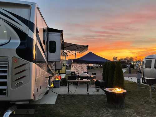 A cozy RV campsite at sunset, featuring a fire pit, outdoor seating, and a blue canopy.