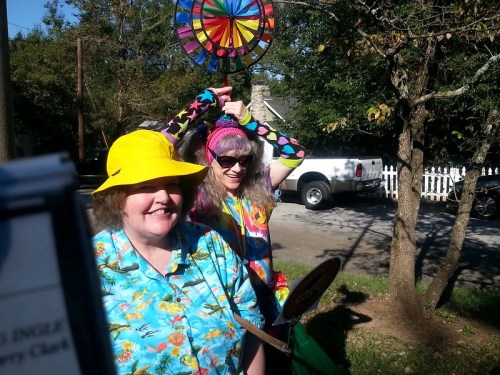 Two women in colorful outfits and hats smile outdoors, with a vibrant wheel decoration in the background.