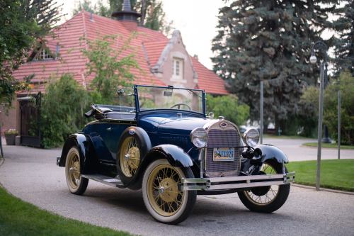 A vintage blue convertible car parked on a scenic driveway with greenery and a historic building in the background.