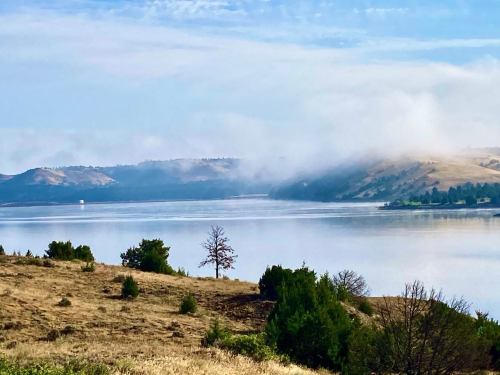 A serene lake surrounded by rolling hills and mist, with sparse trees along the shoreline under a clear blue sky.