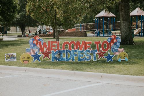 Colorful sign reading "Welcome to Kidfest" with emojis and decorations, set in a park with playground equipment in the background.