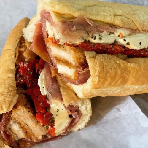 Two hearty sandwiches stacked, filled with layers of meats, cheese, and roasted red peppers, in crusty bread.
